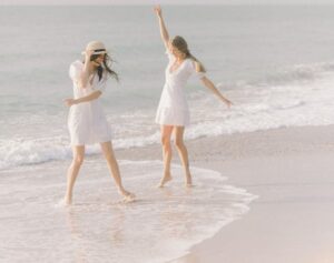 Two women in white dresses enjoying a carefree moment on a sunny beach by the sea.