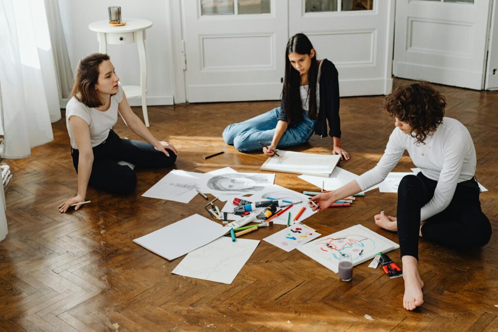 Three women engage in a creative drawing session on a wooden floor, exploring their artistic talents together.