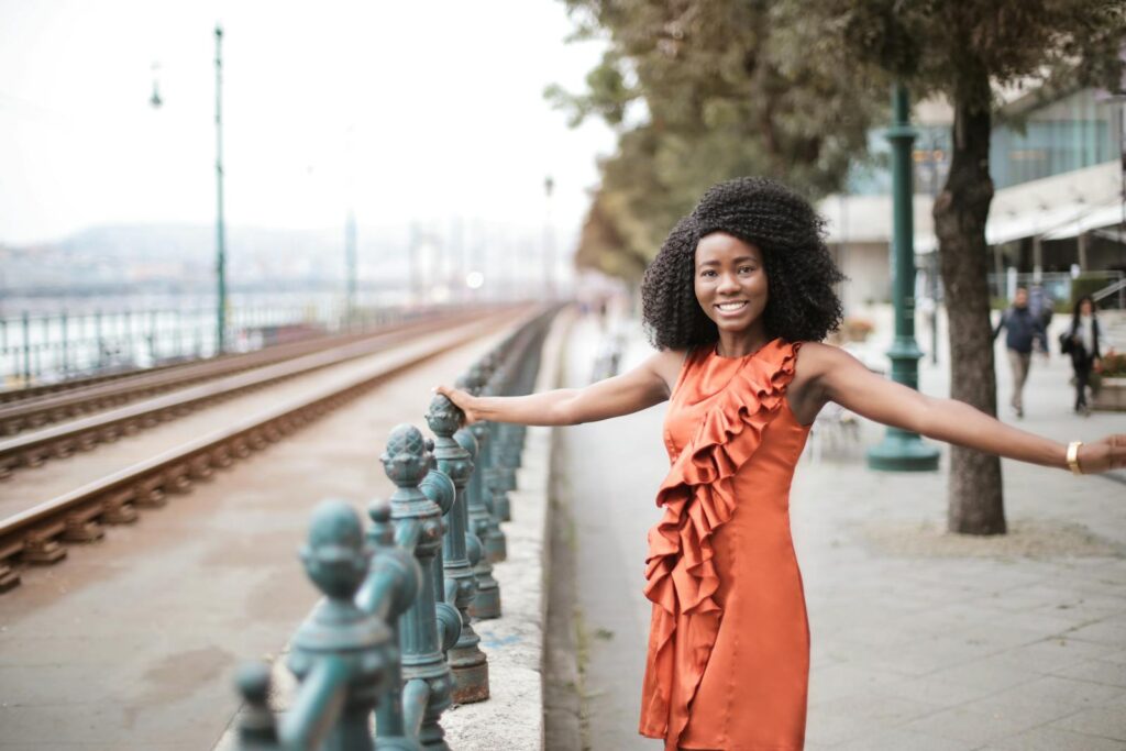 Smiling woman in an orange dress posing joyfully by an urban railway.