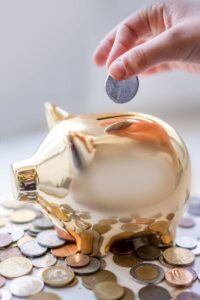 Hand placing coin into a golden piggy bank surrounded by coins, symbolizing savings.
